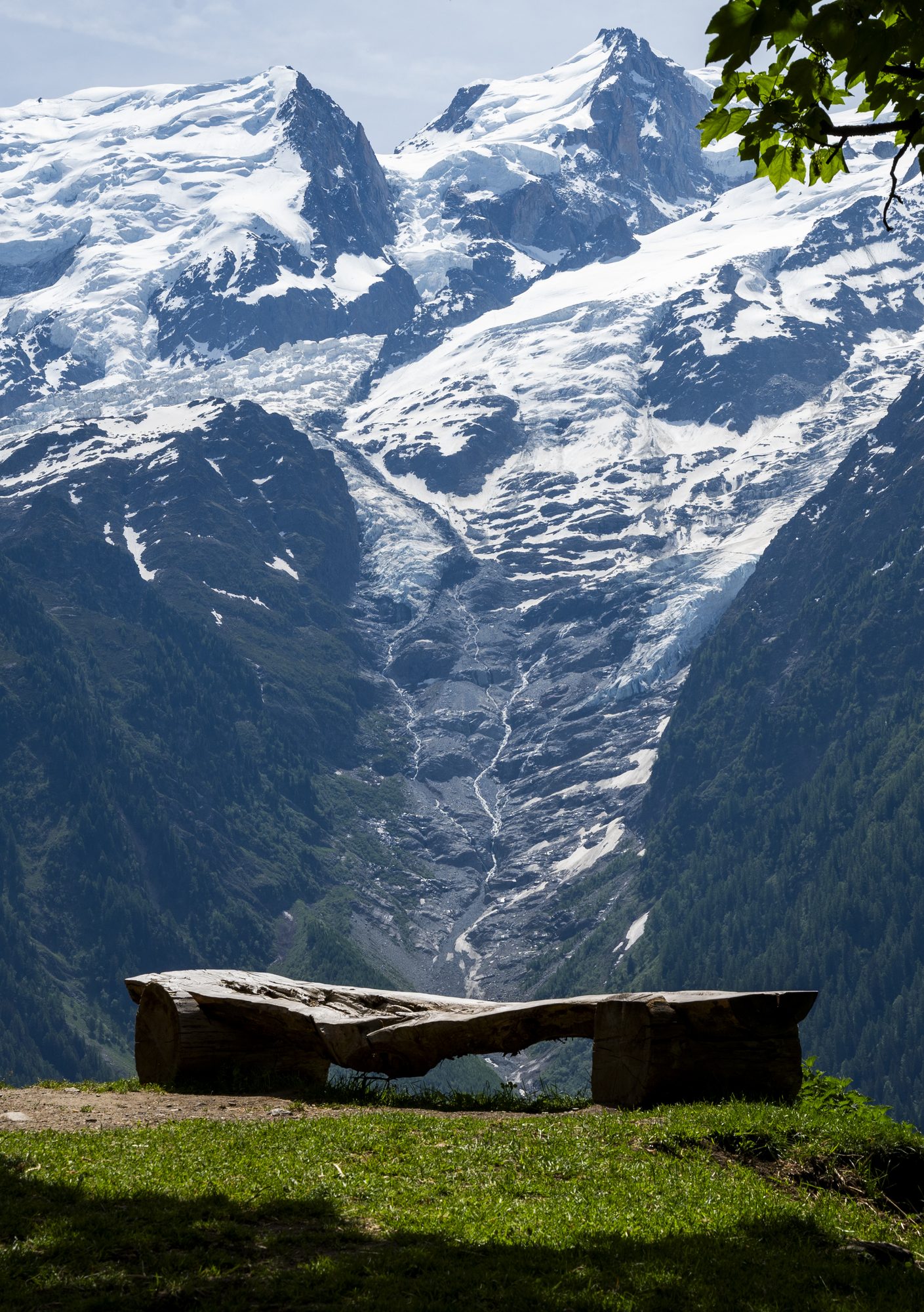 Snow-covered alpine peaks and a glacier valley, with a wooden bench on a grassy ledge in the foreground